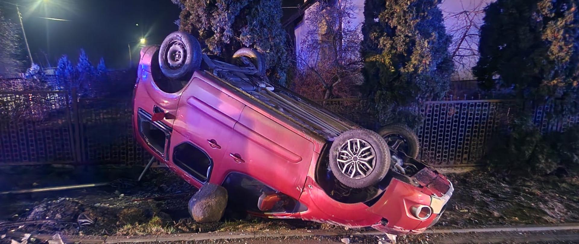 Na zdjęciu widzimy samochód osobowy koloru czerwonego który brał udział w kolizji drogowej. Samochód leży na dachu, liczne rysy i wgniecenia, wybite szyby. Dalej w tle widzimy metalowe ogrodzenie koloru brązowego oraz część budynku mieszkalnego. Pora nocna. 