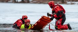 Strażak w kombinezonie wodno-lodowym podaje deskę ortopedyczną do przerębla w której jest strażak udający poszkodowanego