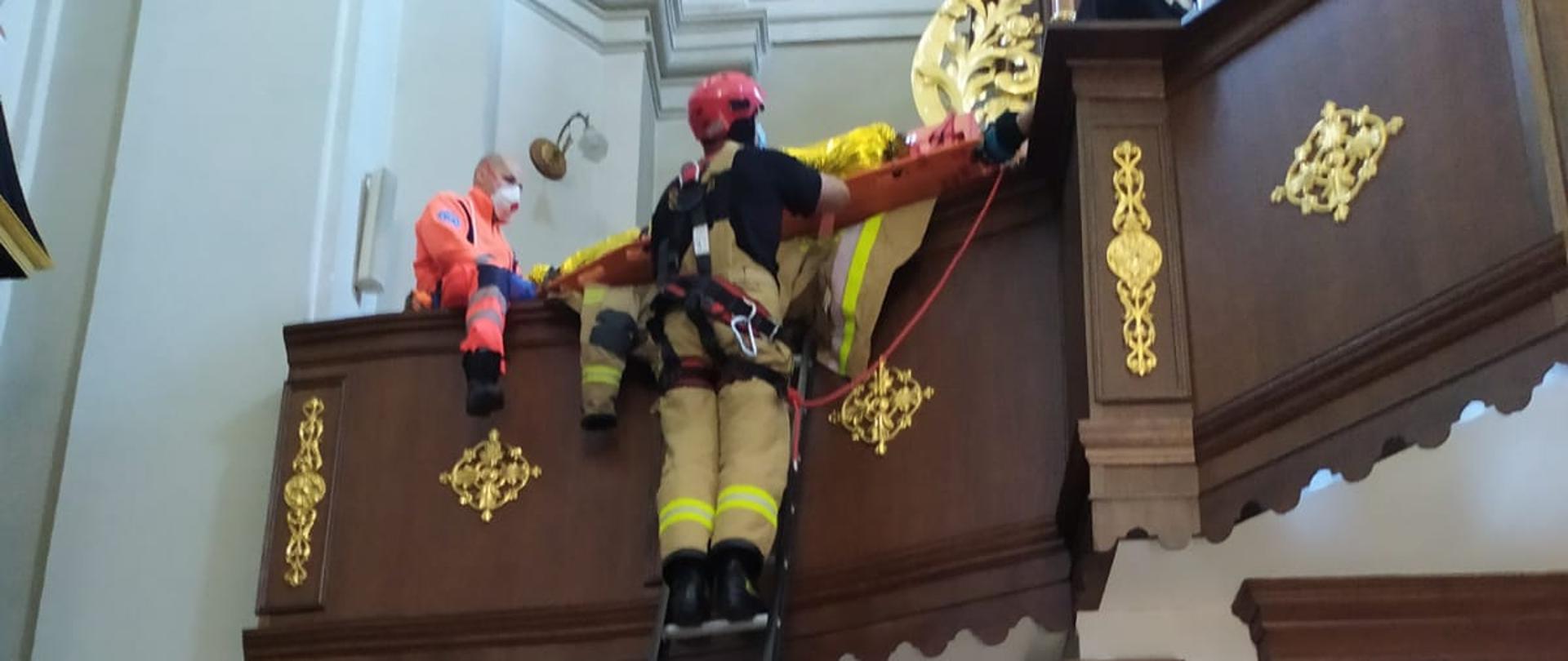 Zdjęcie przedstawia dwóch strażaków trzymających drabinę na której znajduje się trzeci strażak podejmujący poszkodowanego organistę z rąk ratowników z Pogotowia Ratunkowego.