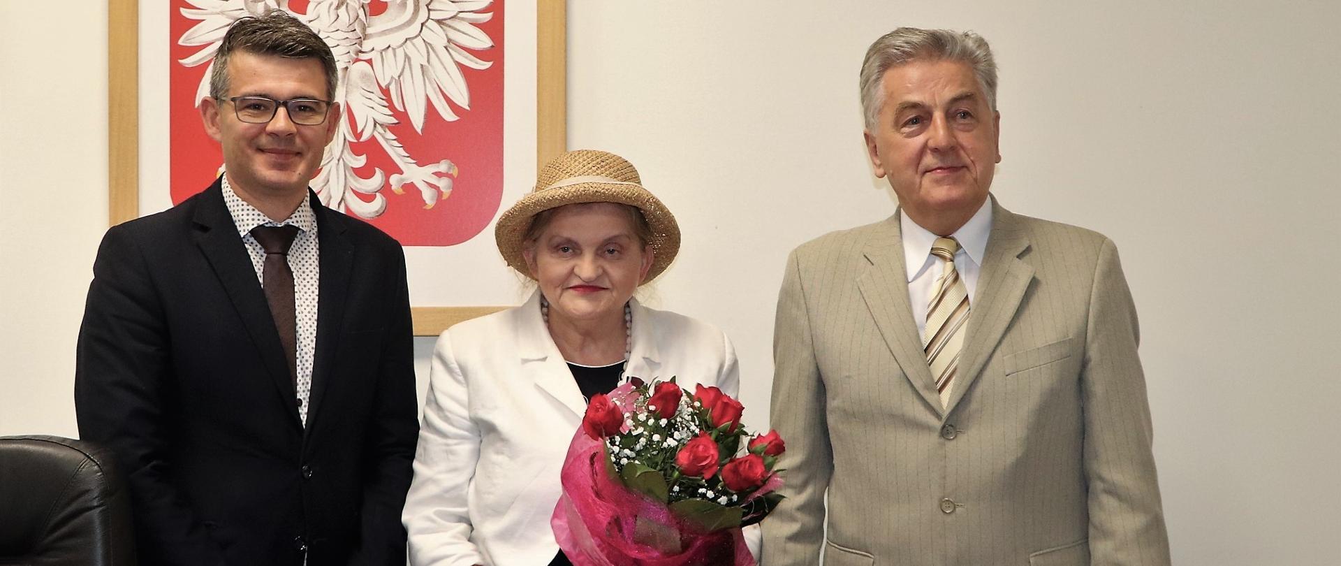Do wspólnego zdjęcia pozuje od lewej - dyrektor Biura Wojewody, odznaczona Aleksandra Sołtysik i wicewojewoda Wojciech Perczak. W tle wisi duże godło w drewnianej ramie 