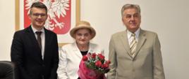 Do wspólnego zdjęcia pozuje od lewej - dyrektor Biura Wojewody, odznaczona Aleksandra Sołtysik i wicewojewoda Wojciech Perczak. W tle wisi duże godło w drewnianej ramie 