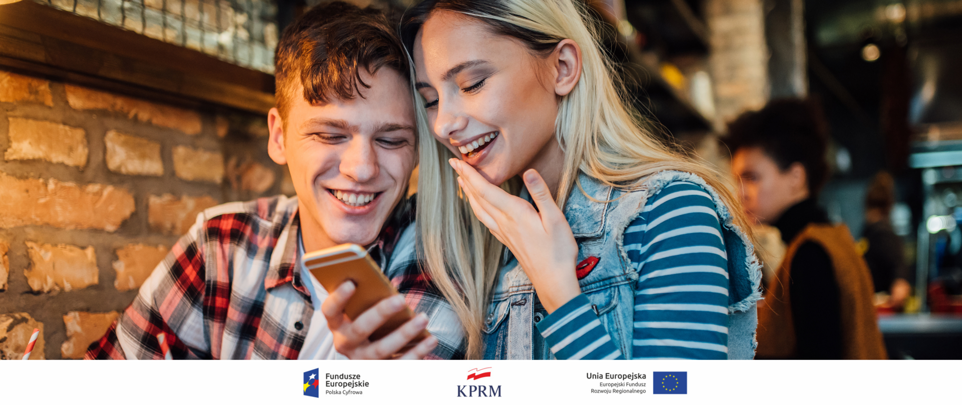 Dwoje uśmiechniętych młodych ludzi (chłopak i dziewczyna) w kawiarni, patrzą w ekran trzymanego przez dziewczynę smartfona.