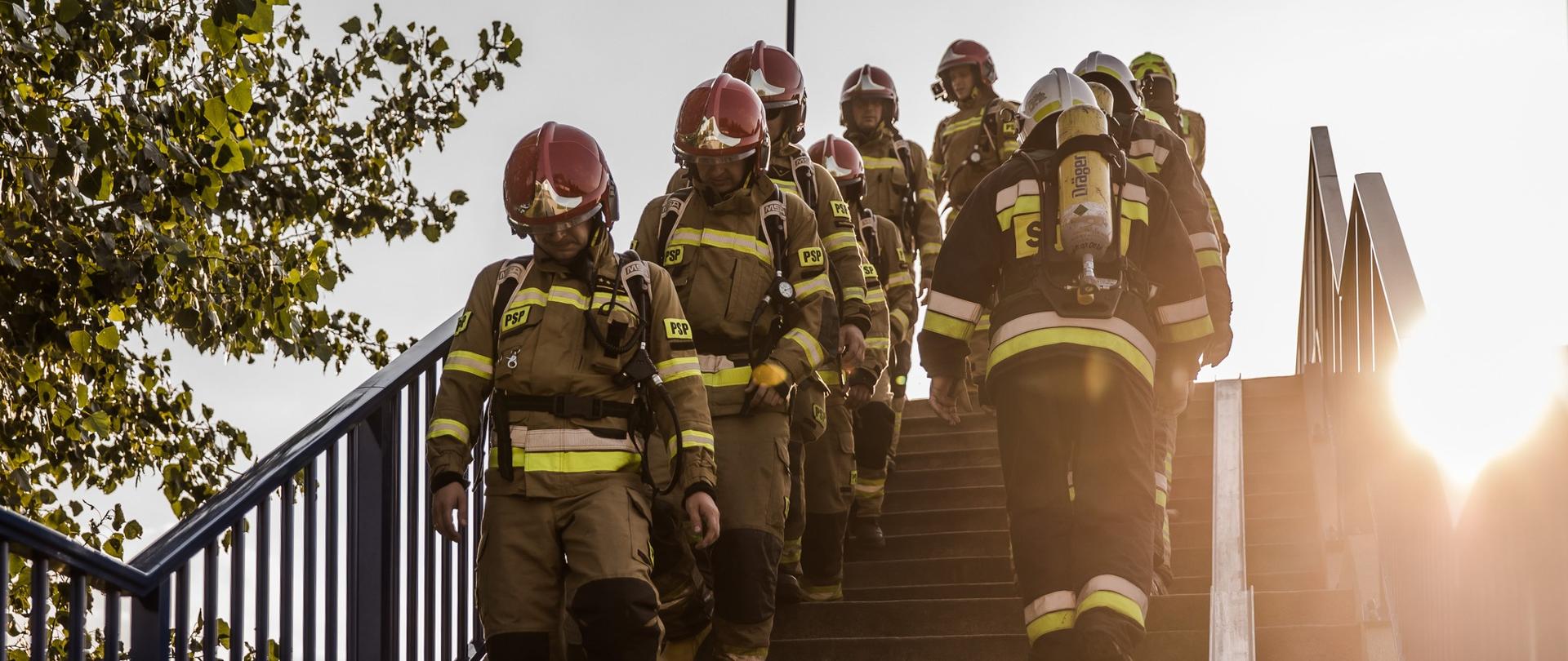 Strażacy w ubraniach specjalnych i z aparatami ochrony dróg oddechowych wchodzą i schodzą po schodach wiaduktu kolejowego.