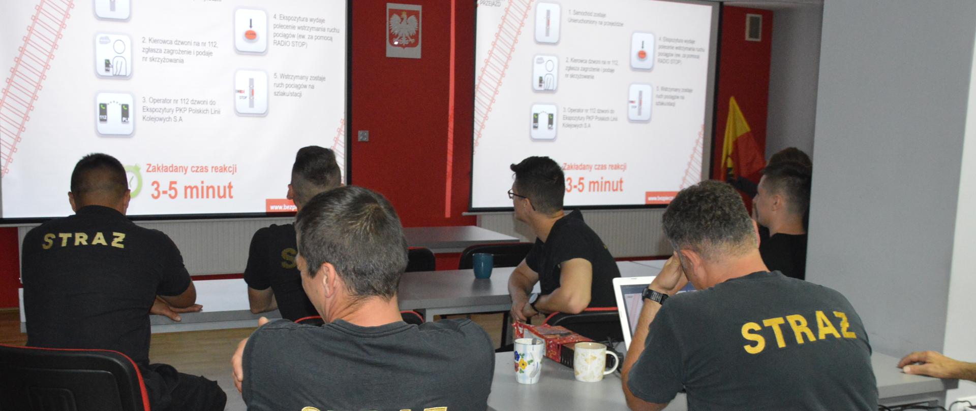 Szkolenie z zakresu bezpieczeństwa na przejazdach kolejowych. Świetlica Komendy Powiatowej PSP w Rawiczu. Przy stołach siedzi 6 strażaków-uczestników szkolenia. Ubrani są w czarne koszulki z krótkim rękawem. Na pierwszym stole znajduje się laptop oraz dwa kubki. W tle - na ekranach wyświetlana jest prezentacja multimedialna. U dołu - czerwony tekst: " Zakładany czas reakcji 3-5 minut".