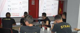 Szkolenie z zakresu bezpieczeństwa na przejazdach kolejowych. Świetlica Komendy Powiatowej PSP w Rawiczu. Przy stołach siedzi 6 strażaków-uczestników szkolenia. Ubrani są w czarne koszulki z krótkim rękawem. Na pierwszym stole znajduje się laptop oraz dwa kubki. W tle - na ekranach wyświetlana jest prezentacja multimedialna. U dołu - czerwony tekst: " Zakładany czas reakcji 3-5 minut".