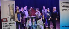 Zdjęcie zbiorowe młodzieży z nauczycielami na scenie po koncercie muzyki dawnej. wszyscy są uśmiechnięci. W tle stoi klawesyn z otwartą klapą. Po lewej stronie stoi baner reklamujący zespół muzyki dawnej, po prawej stronie baner agencji STOART.