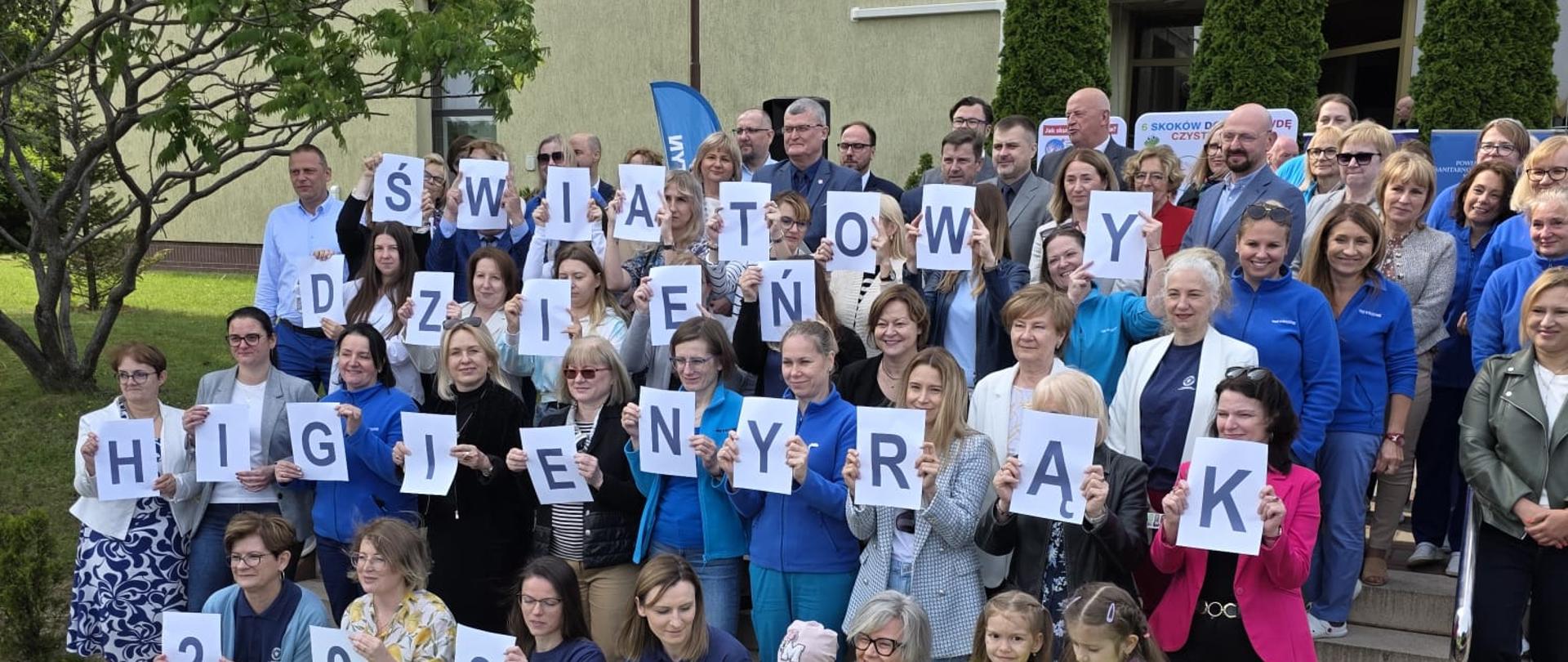 Akcja edukacyjna "Myję ręce, bo wiem więcej". Pamiątkowe zdjęcie organizatorów akcji z dziećmi i zaproszonymi gośćmi, któzy trzymają w dłoniach kartki z literami układającymi się w hasło akcji. 