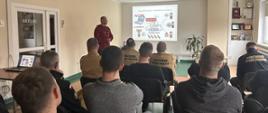 Zdjęcie wykonane na świetlicy KP PSP Białogard. W oddali widać wyświetlaną prezentację poprzez rzutnik. Na tle prezentacji, mężczyzna w czerwonej bluzie, na pierwszym planie widać osoby zgromadzone na sali.Osoby widoczne od tyłu.