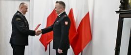 Strażak w umundurowaniu wyjściowym trzyma teczkę i gratuluje strażakowi stojącemu przed nim za nimi ustawione są trzy flagi Polski oraz zegar.