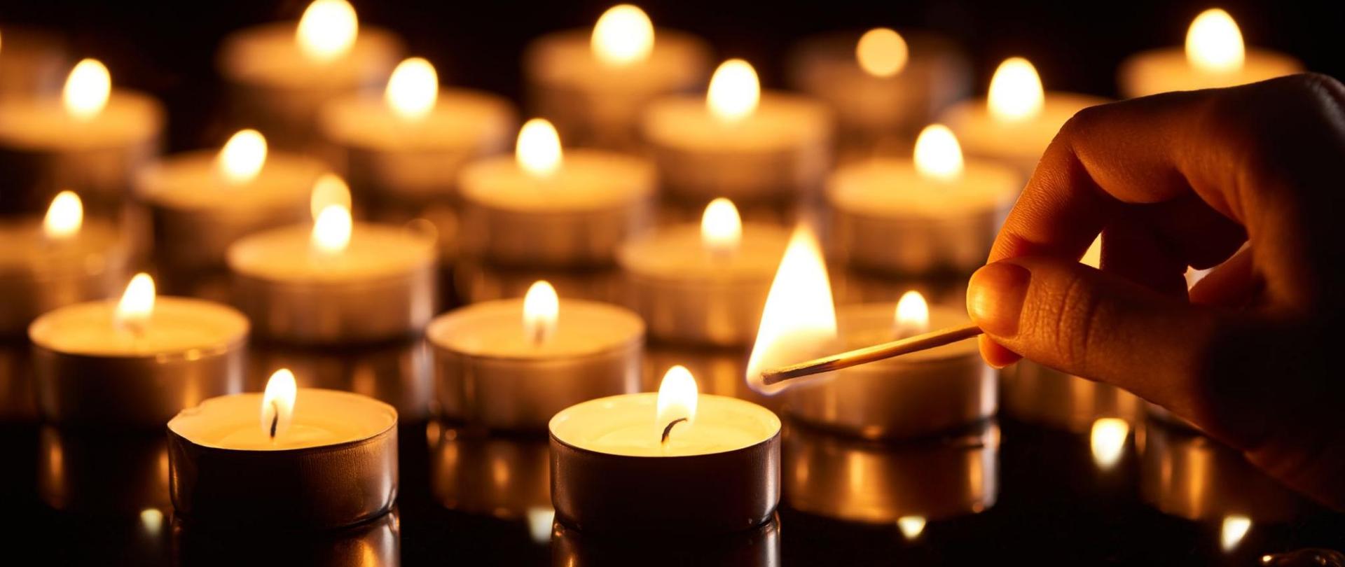 Na zdjęciu znajdują się płonące świeczki. Z prawej strony widać dłoń, w której znajduje się płonąca zapałka.