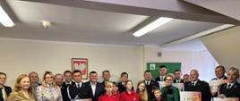 Dwadzieścia trzy osoby mężczyźni i kobiety stoją na tle ściany z godłem polski i banerem KRUS, część z nich w galowych mundurach strażaka trzyma plansze z kwotą otrzymanego wsparcia