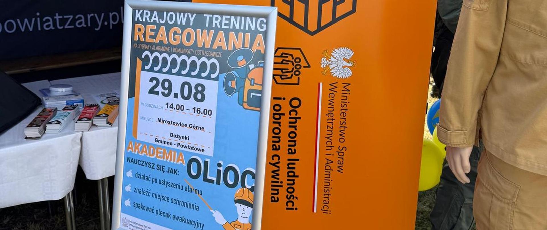 Zbliżenie na materiały informacyjno-edukacyjne przed namiotem. Po prawej stronie stoi pionowy, pomarańczowo-biały roll-up z logo MSWiA, napisem „PROGRAM OLiOC – Ochrona ludności i obrona cywilna” oraz manekin w piaskowym mundurze. Po lewej stronie, na metalowym stojaku, umieszczony jest plakat informacyjny z napisem „KRAJOWY TRENING REAGOWANIA – 29.08, godz. 14.00–16.00, Mirostowice Górne, Dożynki Gminno-Powiatowe”.
