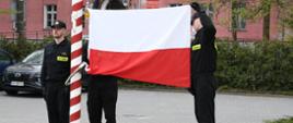 Strażacy stoją przy maszcie i uroczyście wieszają flagę Polski.