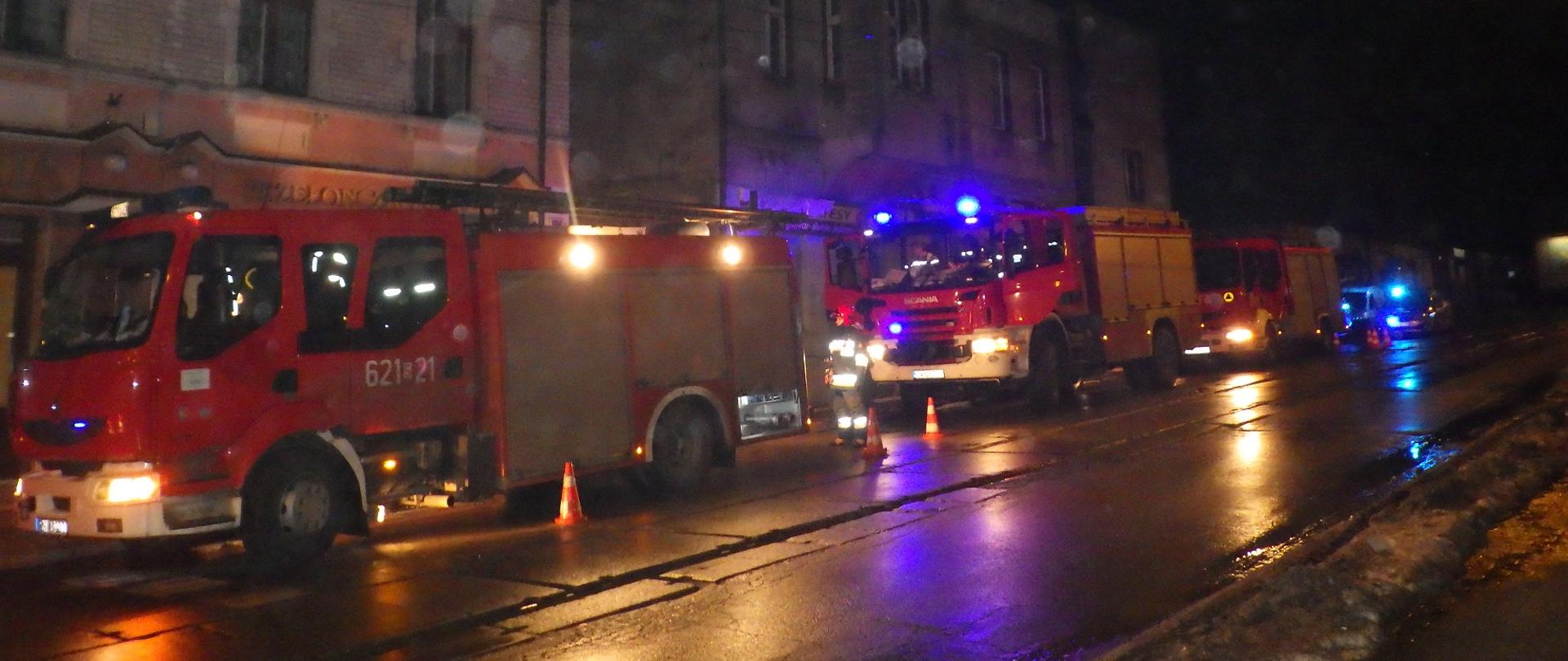 Zdjęcie przedstawia samochody pożarnicze stojące na ulicy w pobliżu budynku, w których wybuchł pożar. Zdjęcie wykonane w nocy.