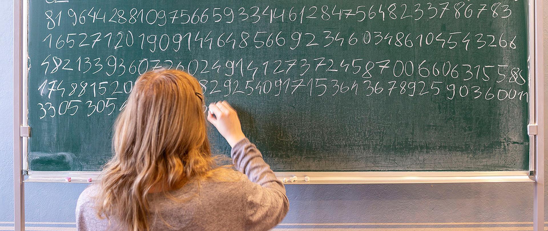 Zbliżenie na dużą, zieloną tablicę szkolną, która jest niemal w całości zapisana białym kreda. Widzimy dużo kolejnych cyfr po przecinku. Odwrócona plecami dziewczyna o jasnych włosach właśnie dopisuje kolejną cyfrę w dolnym rzędzie.