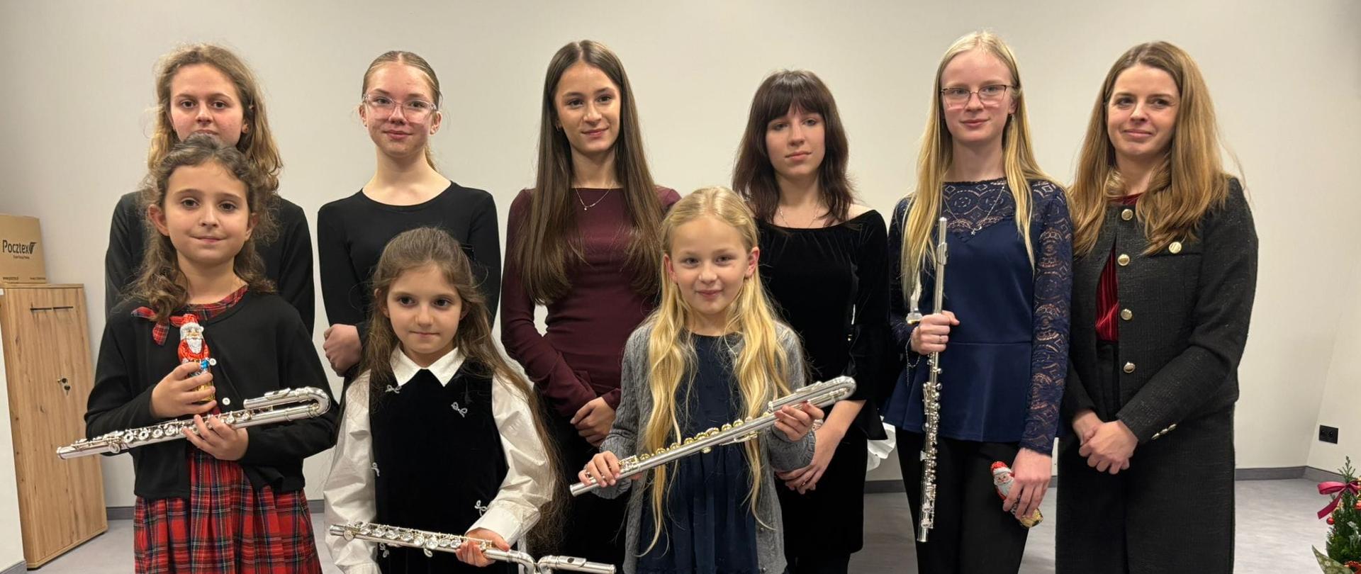 Na zdjęciu widać grupę dziewięciu flecistek w różnym wieku oraz jedną dorosłą osobę – nauczycielkę. Wszyscy stoją w jasnej, przestronnej sali o gładkich, jasnych ścianach i szarej podłodze. Dziewczęta ustawione są w dwóch rzędach: młodsze z przodu, starsze z tyłu. Większość trzyma srebrne flety poprzeczne; kilka osób ma w dłoniach małe figurki świąteczne, co sugeruje grudniowy, świąteczny charakter wydarzenia. Uczennice są ubrane elegancko – dominują ciemne sukienki, spódnice i bluzki, niektóre z delikatnymi koronkami. Młodsze dziewczynki mają rajstopy i lakierowane buty. Nauczycielka stoi z boku grupy, ubrana elegancko, z ciepłym, spokojnym wyrazem twarzy. Cała scena sprawia wrażenie uroczystej, ale bardzo serdecznej.