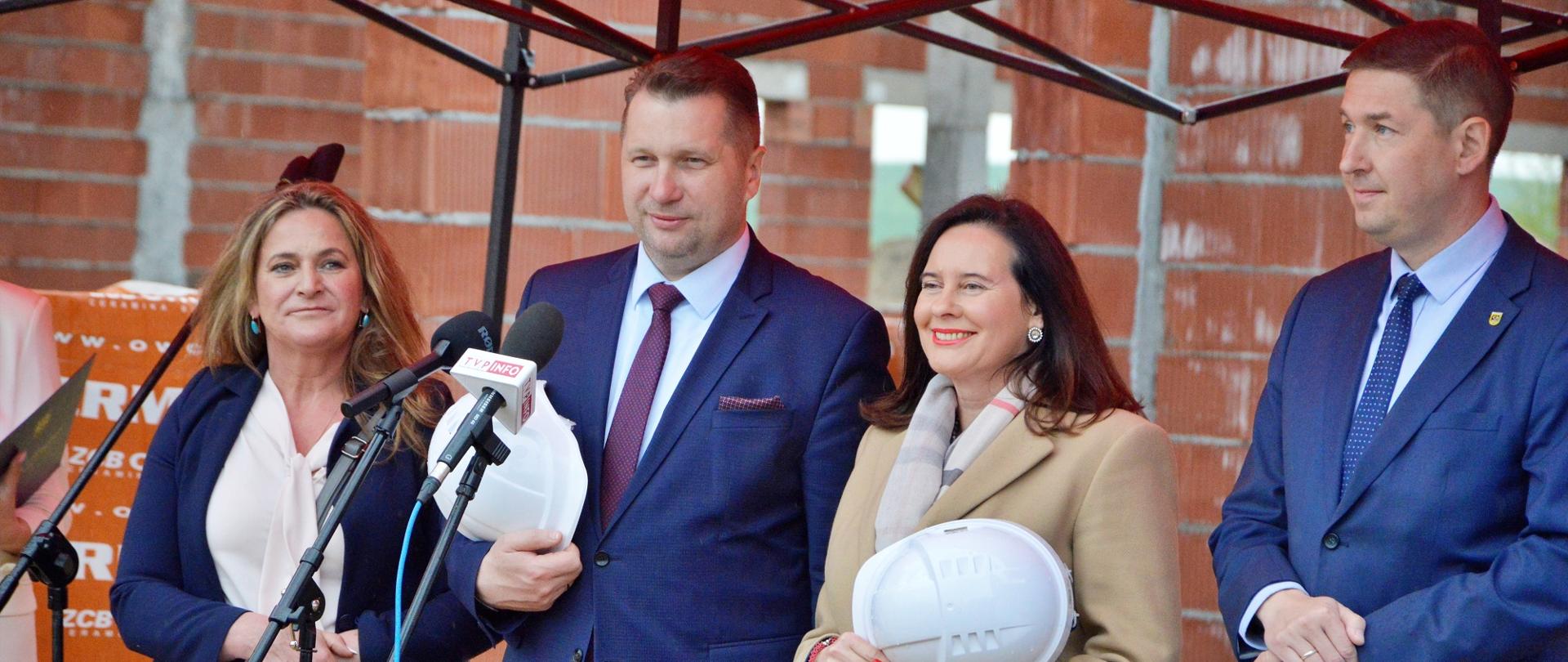 Na placu budowy stoją: kobieta w granatowej marynarce, minister Czarnek, kobieta w jasnym szarożółtym płaszczu i mężczyzna w garniturze, wszyscy trzymają w rękach białe kaski, minister mów do kilku mikrofonów na stojakach, za nimi widać ściany z pomarańczowych pustaków i rusztowania.