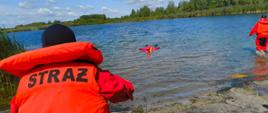 Widok znad brzegu na scenę ratunkową na jeziorze. Na pierwszym planie, z tyłu, widać ratownika w czerwonym kombinezonie i kamizelce ratunkowej, z napisem "STRAŻ" na plecach, trzymającego linę. Lina prowadzi w kierunku środka jeziora, gdzie inny ratownik w czerwonym kombinezonie unosi się na wodzie, trzymając się pomarańczowej boi ratunkowej. W oddali, po prawej stronie, w wodzie stoi trzeci ratownik, również w czerwonym kombinezonie. Brzegi jeziora są porośnięte roślinnością i drzewami. Nad jeziorem widać błękitne niebo z białymi, puszystymi chmurami.