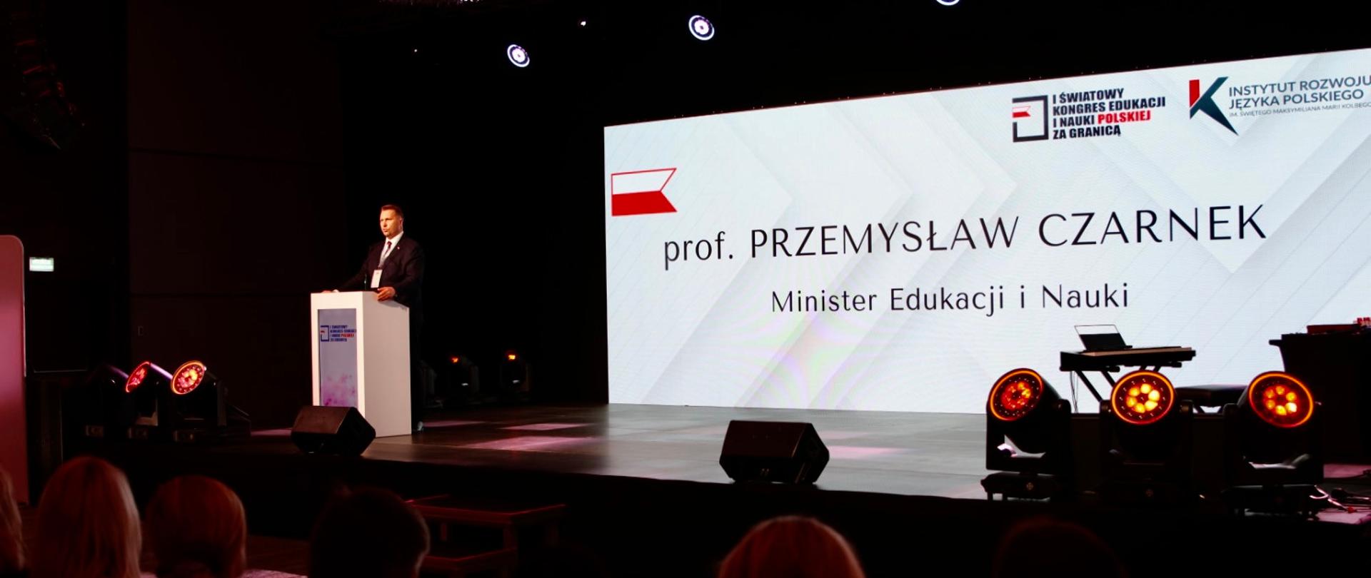 Za mównicą stoi minister Czarnek i mówi do mikrofonu, za nim na ścianie napis Prof. Przemysław Czarnek, Minister Edukacji i Nauki, przed nim zaciemniona sala.