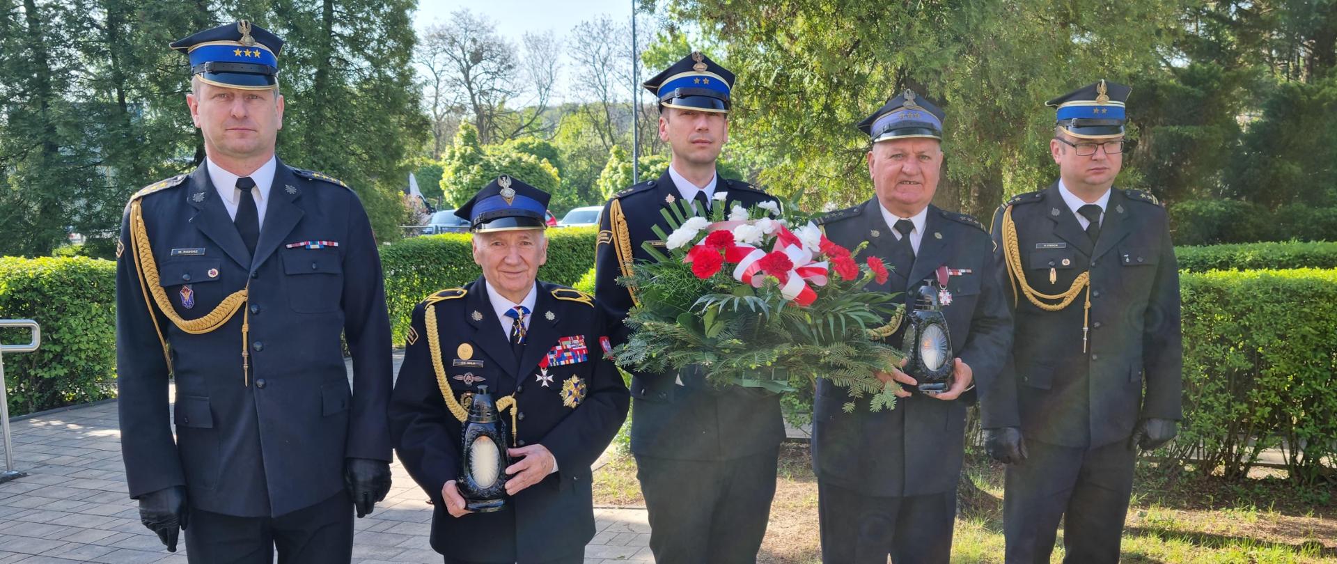 Na zdjęciu Komendant Miejski Państwowej Straży Pożarnej starszy brygadier Marek Radosz wraz z Zastępcami brygadierem Kamilem Dzwonnikiem oraz brygadierem Pawłem Frochem, z którymi stoją w rzędzie prezesi kół związku emerytów i rencistów pożarnictwa działających przy Komendzie Miejskiej Państwowej Straży Pożarnej w Częstochowie. Zastępca Komendanta brygadier Kamil Dzwonnik stojący w środku trzyma wiązankę prezesi kół emerytów stoją po jego obu stronach trzymając znicze po zewnętrznych stronach rzędu stoi Komendant Miejski Państwowej Straży Pożarnej starszy brygadier Marek Radosz oraz Zastępca Komendanta Miejskiego brygadier Paweł Froch. W tle widać chodnik, żywopłot oraz drzewa. Pogoda jest słoneczna, a atmosfera sugeruje powagę i zamyślenie. Wszyscy ubrani w mundury wyjściowe.