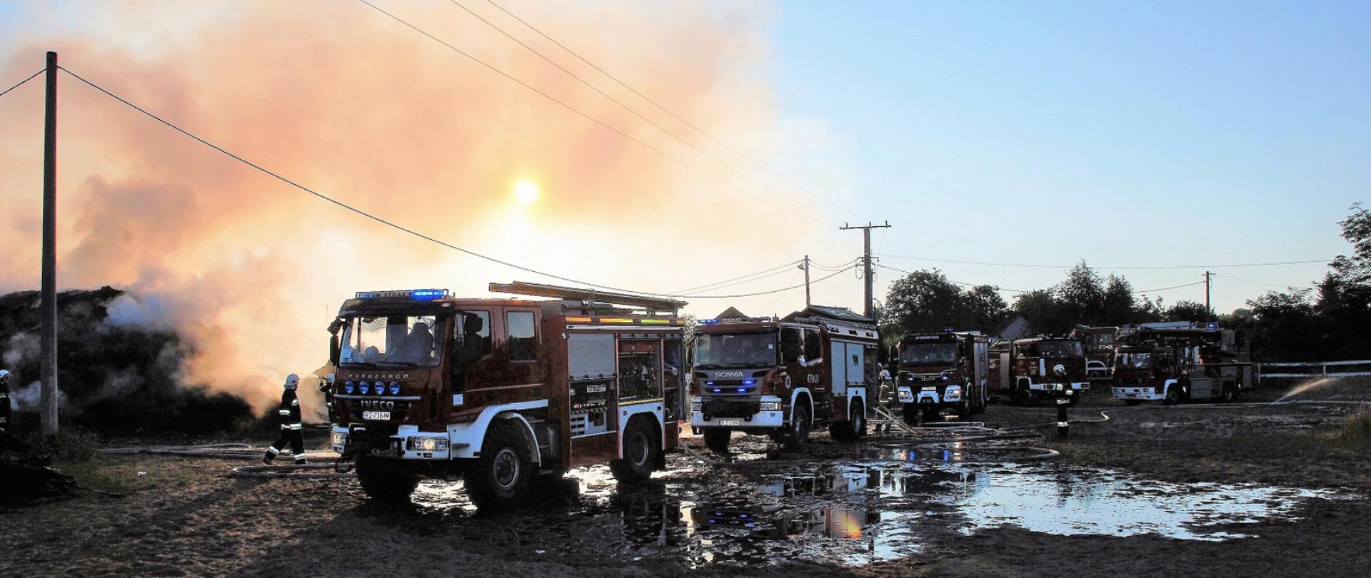 Na zdjęciu widzimy sześć samochodów pożarniczych ciężarowych z których są rozwinięte linie gaśnicze, widać chodzących strażaków w pełnym uzbrojeniu. W tle widać dymiące się sterty gaszonego siana.