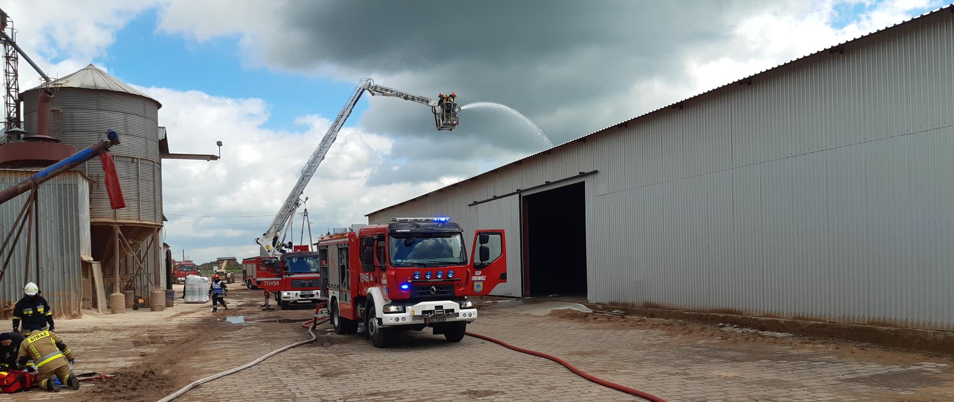 Dwa samochody ratowniczo- gaśnicze straży pożarnej stoją miedzy halami produkcyjnymi peletu. Z tyłu z podnośnika strażacy leją wodę na jedną z hal. Po prawej stronie zdjęcia strażacy klęczą z torbą ratowniczą R1.