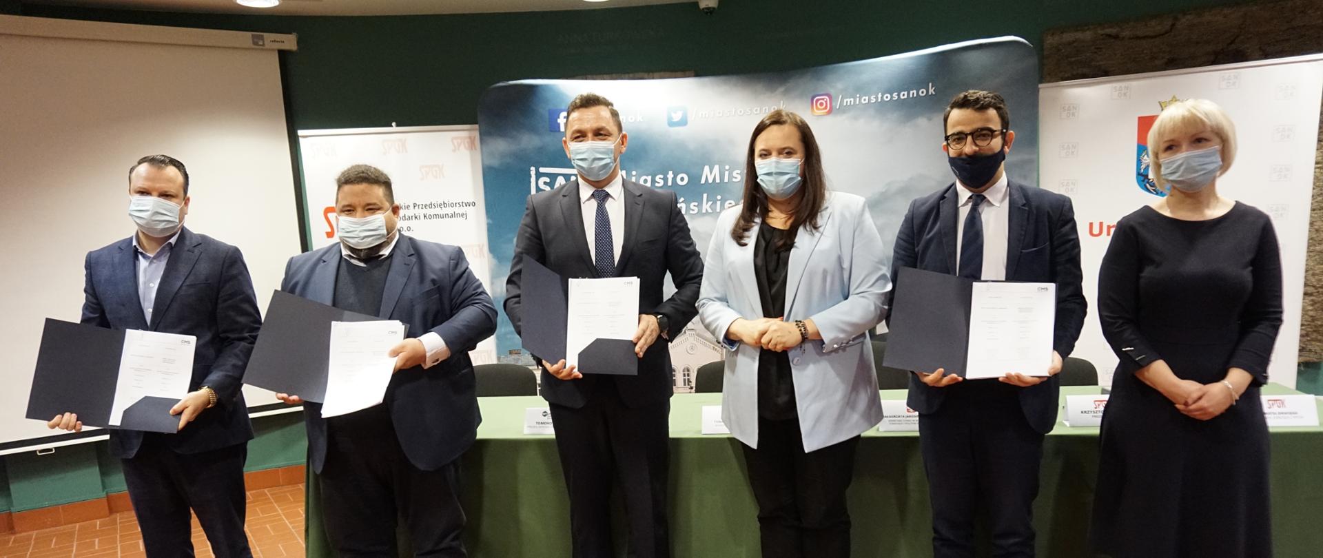 Na zdjęciu stoi sześć osób, trzecia od prawej - wiceminister Małgorzata Jarosińska-Jedynak, prezentują podpisane dokumenty, w tle bannery reklamowe Miasta Sanok i stół konferencyjny