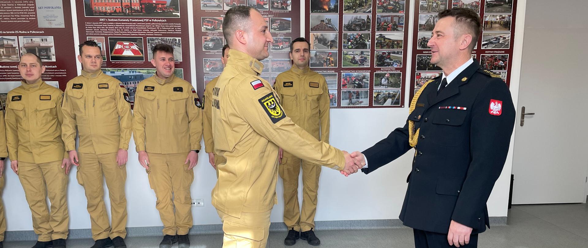 Na zdjęciu przedstawiona jest oficjalna uroczystość wewnątrz jednostki Państwowej Straży Pożarnej (PSP).
---
## Opis pierwszego planu
* **Mężczyzna po prawej stronie:** Stoi profilem, ubrany w wyjściowy, ciemnogranatowy lub czarny mundur strażacki. Na lewym ramieniu ma naszywkę z godłem Polski (orłem), na piersi baretki odznaczeń, a przy ramieniu złoty sznur galowy (akszlevant). Podaje rękę stojącemu naprzeciwko strażakowi.
* **Mężczyzna w centrum:** Stoi profilem, ubrany w piaskowy (beżowy) mundurowy strój służbowy PSP. Na ramieniu ma naszywkę z polską flagą oraz okrągły emblemat jednostki dolnośląskiej z czarnym orłem. Wymienia uścisk dłoni z oficerem w ciemnym mundurze.
---
## Opis drugiego planu i tła
* **Grupa strażaków:** W tle, pod ścianą, stoi w szeregu czterech innych strażaków ubranych w identyczne, piaskowe mundury służbowe. Obserwują oni ceremonię uścisku dłoni.
* **Tablice informacyjne:** Na ścianie za strażakami wiszą duże, bordowe tablice pamiątkowe lub informacyjne z nagłówkami zawierającymi liczbę „30”. Znajdują się na nich liczne zdjęcia dokumentujące historię jednostki, opisy (w tym tekst dotyczący Komendy Powiatowej PSP w Polkowicach) oraz fotografie operacyjne z akcji ratowniczych.
* **Pomieszczenie:** Podłoga jest jasnoszara, gładka, a po prawej stronie w tle widoczne są białe drzwi wejściowe. Na podłogę wpada jasne światło dzienne.