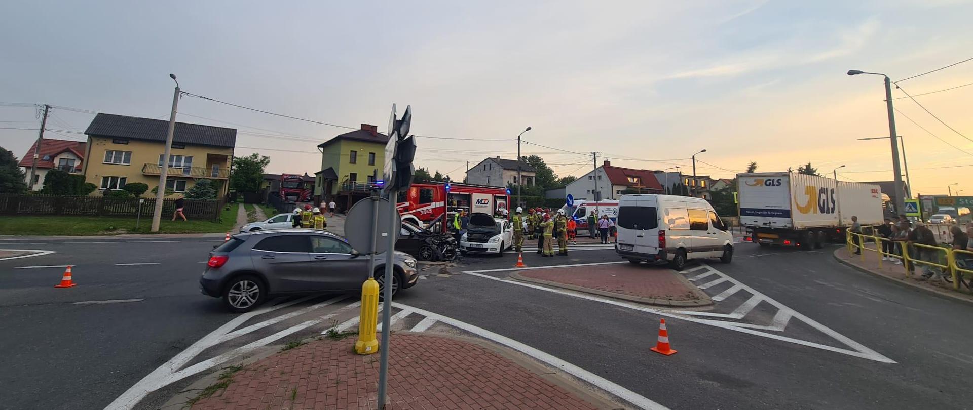 Zdjęcie przedstawia skrzyżowanie, na którym doszło do zderzenia dwóch samochodów osobowych. Ruch odbywa się jednym awaryjnym pasem, za którym widać dwa rozbite auta. W dalszej perspektywie stoją samochody pożarnicze i ambulans.