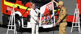 Widok z przodu: Minister klimatu i środowiska Pani Anna Moskwa oraz komendant główny Państwowej Straży Pożarnej gen. brygadier Andrzej Bartkowiak prezentują plakat promujący program "Bitwa o remizy 2023". W tle wozy strażackie. 