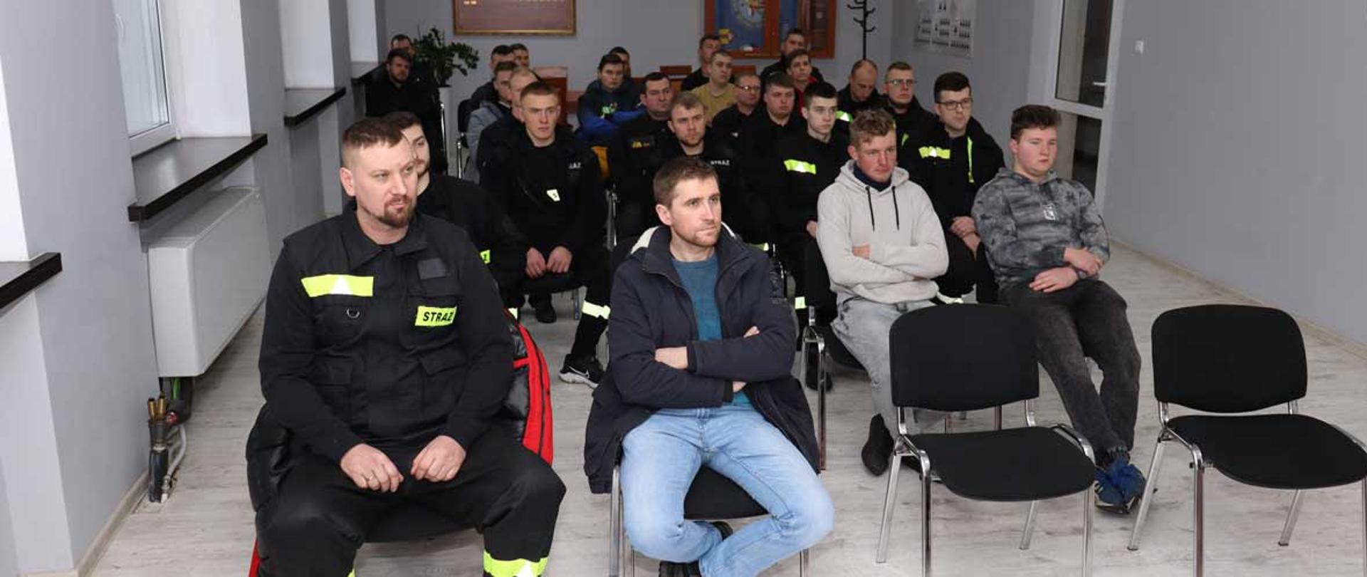 kilkanaście osób ubranych w ubrania koszarowe ochotniczej straży pożarnej barwy czarnej oraz ubrania cywilne siedzi na krzesełkach i słucha przemówienia zastępcy komendanta powiatowego psp w Łowiczu brygadiera Rafał Garbarza w tle wisząca szafka wieszak na ubrania sztandar w drewnianej gablocie po lewej stronie widoczne cztery okna po prawej stronie w środkowej części ściany drzwi plastikowe barwy białej na pierwszym planie w prawym rogi widoczne dwa puste krzesła z czarnym materiałowym obiciem na podłodze panele barwy jasnoszarej