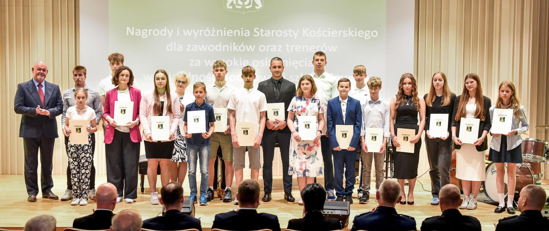 Na zdjęciu grupa młodzieży. Wszyscy odświętnie ubrani. Każdy trzyma teczkę z logo powiatu kościerskiego. W tyle za nimi na ścianie napis "nagrody i wyróżnienia Starosty Kościerskiego dla zawodników i trenerów za wysokie osiągnięcia w działalności sportowej".