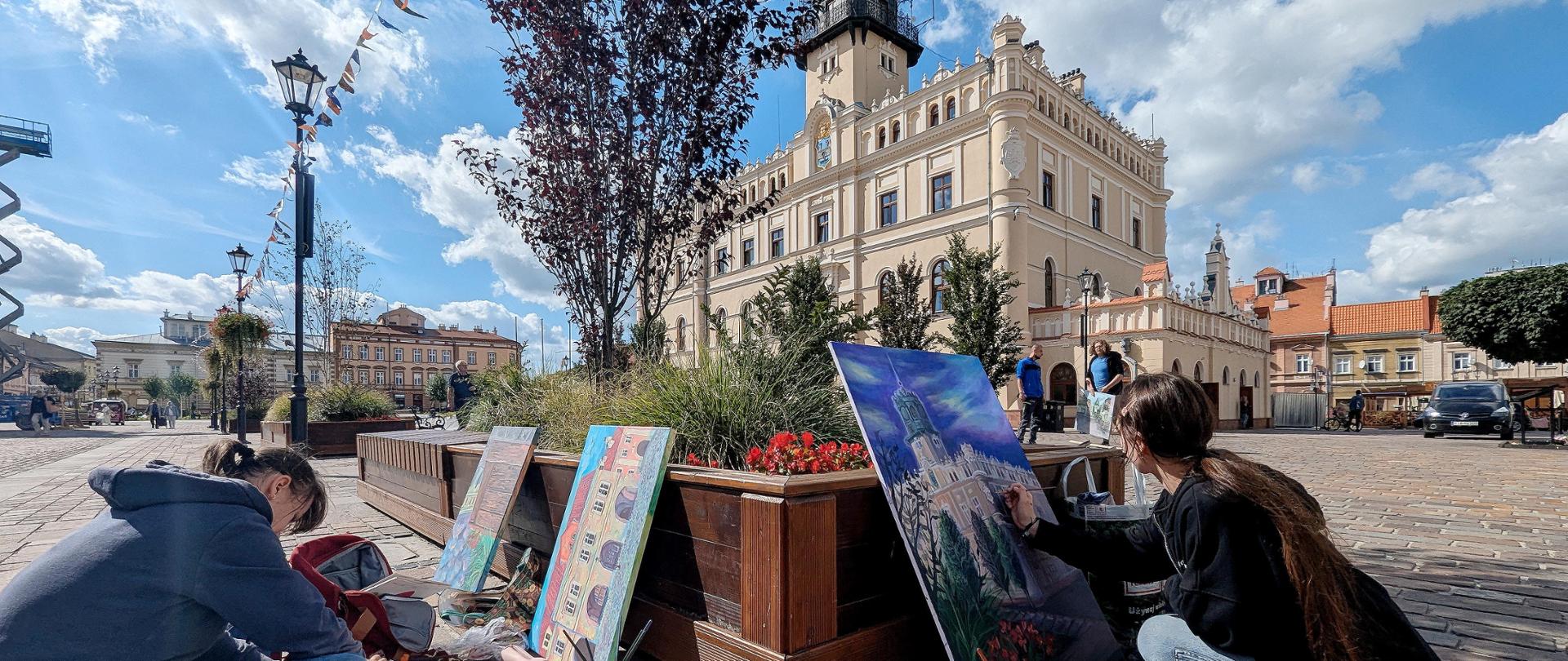 Na pierwszym planie dwie kobiety malują obrazy. W tle widać budynek ratusza, który jest tematem jednej z prac.