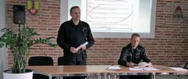 strażak ubrany w ubranie sztabowe stoi przed stolikiem, trzyma w dłoni zwiniętą kartkę, drugi strażak ubrany w ubranie sztabowe siedzi za stolikiem i spogląda w otwarty zeszyt, na stoliku leżą dokumenty, za ich plecami ściana z cegły i ekran projekcyjny na którym wyświetla się strona internetowa z zagadnieniami szkoleniowymi strażaków osp, na pierwszym planie stoi duża donica z rośliną o drobnych liściach, na ścianie za nimi po bokach ekranu widoczny herb powiatu łowickiego oraz logo Państwowej Straży Pożarnej