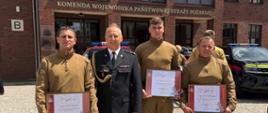 Na zdjęciu widać czterech mężczyzn w mundurach strażackich, którzy stoją na brukowanym placu przed dużym ceglanym budynkiem. Nad wejściem do budynku widnieje napis: "Komenda Wojewódzka Państwowej Straży Pożarnej". Trzech strażaków w piaskowych mundurach polowych trzyma w rękach oprawione dyplomy w ramkach, na których nagłówek zaczyna się od słowa "DYPLOM". Czwarty mężczyzna, pośrodku grupy, jest ubrany w ciemny, galowy mundur z odznaczeniami. W tle widać zaparkowane wozów bojowych straży pożarnej. Zdjęcie jest oświetlone mocnym słońcem, rzucającym wyraźne cienie.