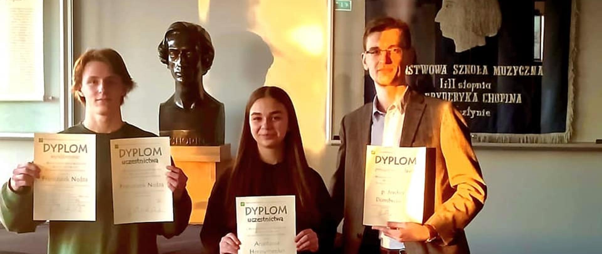 Dwoje uczniów i nauczyciel z dyplomami uczestnictwa w Państwowej Szkole Muzycznej w Olsztynie podczas Makroregionalnego Konkursu z Kształcenia Słuchu