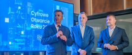 Pod wielkim niebieskim ekranem z napisem Cyfrowe Otwarcie Roku 2024 stoi minister Wieczorek który mówi do trzymanego w ręku mikrofonu i dwóch mężczyzn w garniturach.