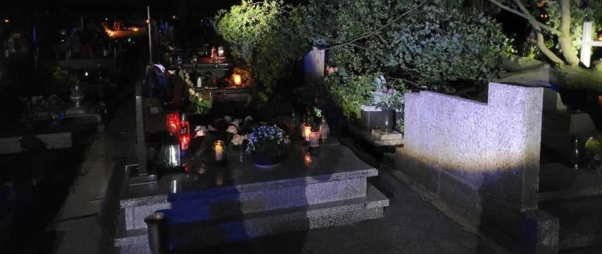 Zdjęcie przedstawia fragment cmentarza w Zielonej Wsi w porze nocnej. Widoczne są nagrobki, na których znajdują się kwiaty oraz zapalone znicze. Z prawej strony widać dużą koronę drzewa i fragment pnia, które leżą na pomnikach. 