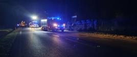 Kolizja na drodze krajowej. Auto ciężarowe leży w rowie na swoim prawym boku. Wokół rozsypany jest materiał przewożony pojazdem (kruszywo). Na jezdni stoją wozy straży pożarnej i radiowóz policyjny. Wszystkie pojazdy mają włączone niebieskie światła błyskowe.