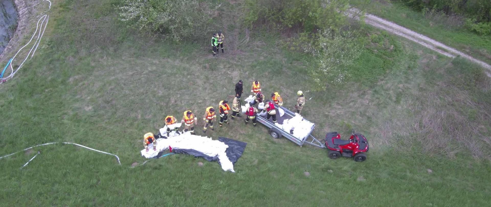 Strażacy układający worki z piaskiem wokół przesiąku, w tle quad i drzewa