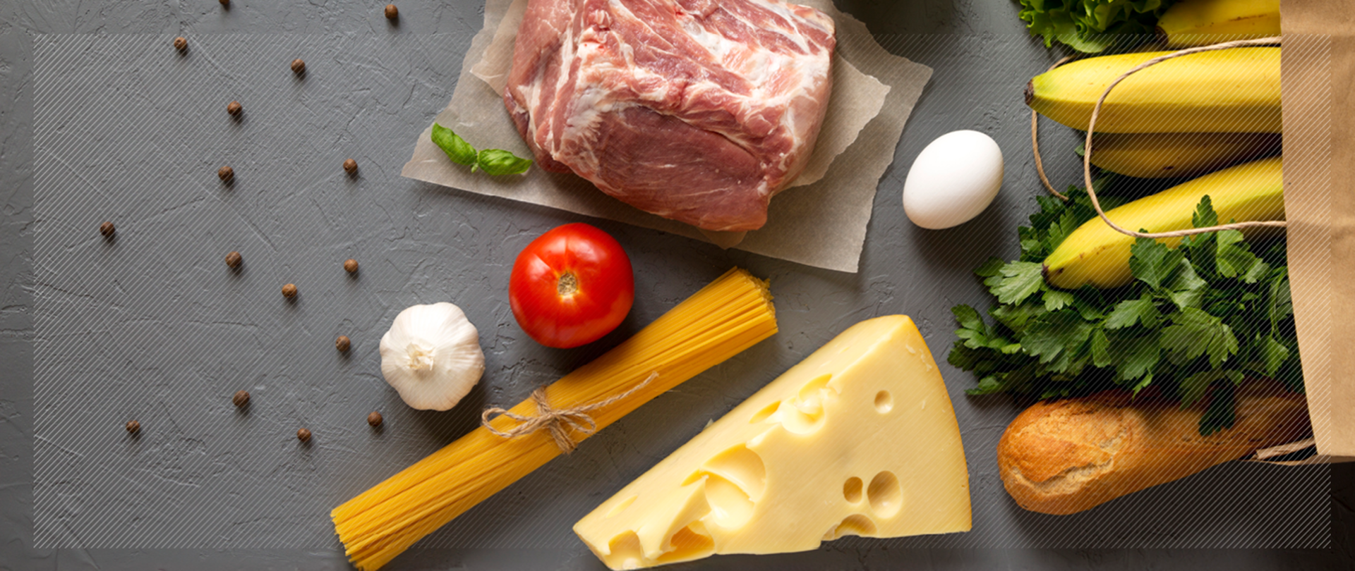 Na szarym blacie leżą warzywa i owoce, ser, mięso, jajka, makaron, bagietka