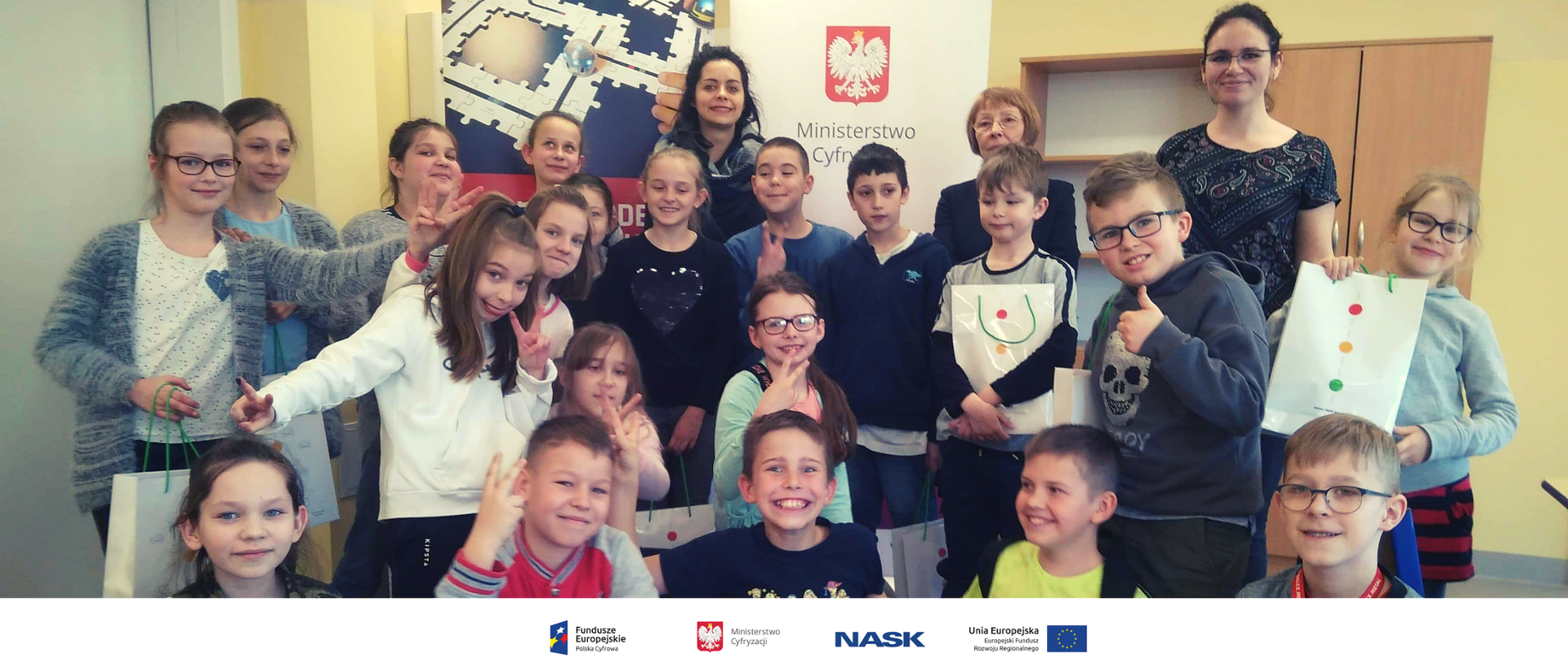 Na zdjęciu widać grupę dzieci w szkolnej klasie. Na dole zdjęcia umieszczone są logotypy: Fundusze Europejskie. Polska Cyfrowa, Ministerstwo Cyfryzacji, NASK oraz Unia Europejska. Europejski Fundusz Rozwoju Regionalnego. 