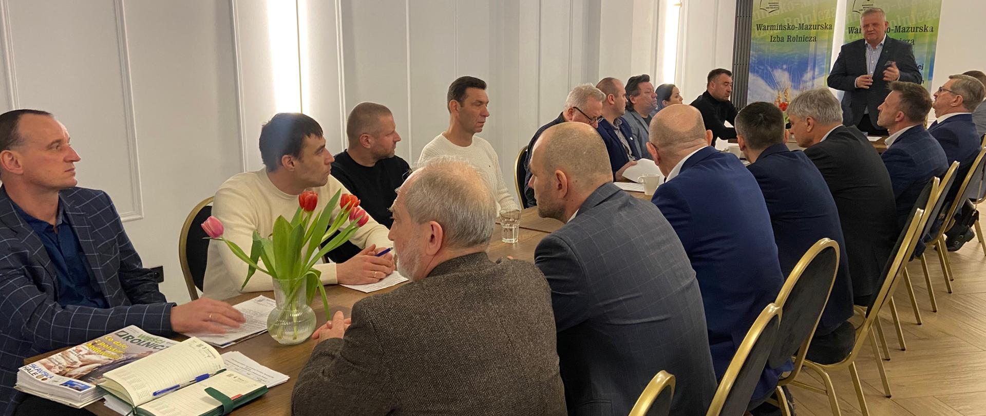 Grupa osób siedzi po obu stronach stołu i zwrócona jest w stronę stojącego w głębi zdjęcia prowadzącego. Za jego plecami banery WMIR 