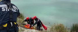 Na tle wody i skarpy widać dwóch strażaków z specjalistycznej grupy ratownictwa wysokościowego, który zabezpieczeni linami ewakuują po skarpie po poszkodowanego. W tle widać jezioro.