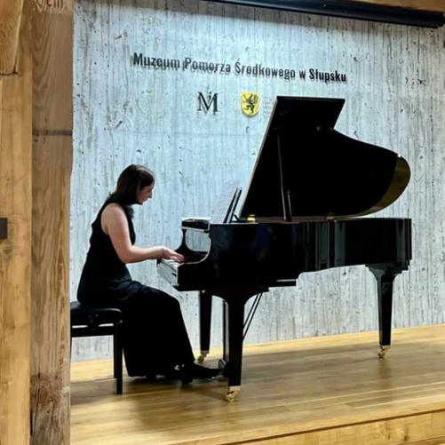 Zdjęcie kolorowe. Młoda pianistka w czarnej sukni gra na fortepianie. Drewniana scena. W tle napis Muzeum Pomorza Środkowego.