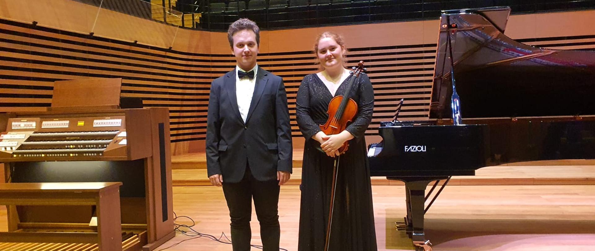Mężczyzna i kobieta stoją na estradzie, kobieta trzyma przed sobą skrzypce i smyczek, po prawej stronie stoi fortepian Fazioli, po lewej stronie stoją organy.