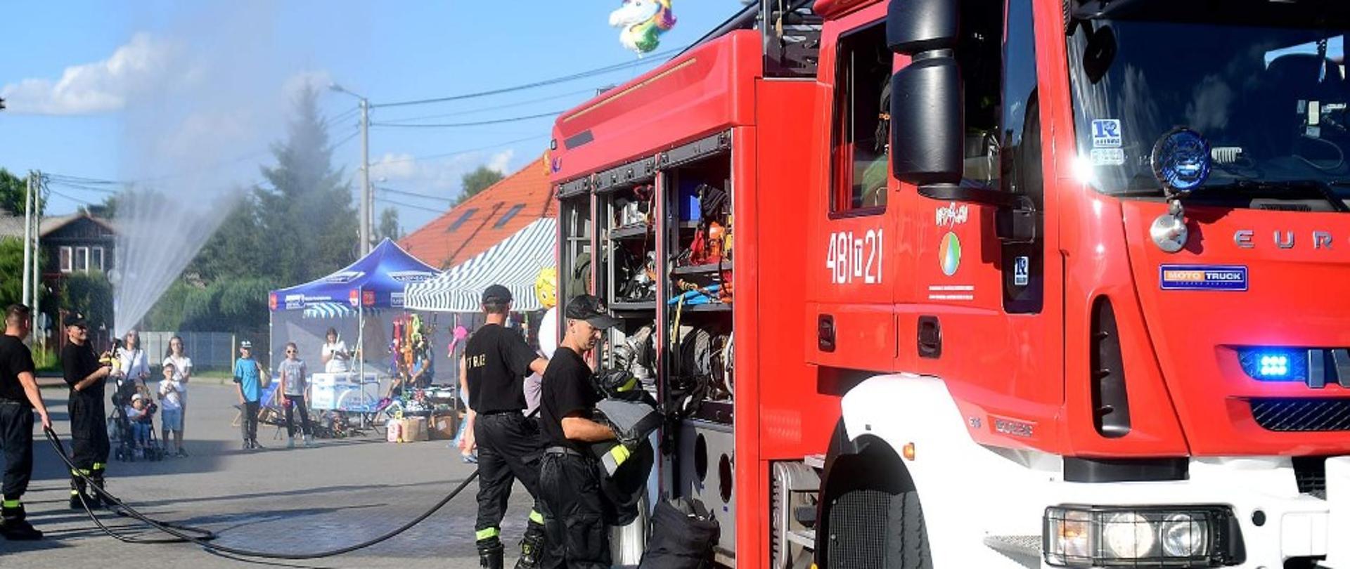 Na pierwszym planie z prawej strony widoczny jest czerwony pojazd gaśniczy Państwowej Straży Pożarnej. W samochodzie za kabiną kierowcy w części składowania sprzętu ratowniczo gaśniczego otworzone są żaluzje i widoczny jest tam zamocowany sprzęt. Przy pojeżdzie stoi dwóch ratowników. Nieco dalej z lewi strony również widoczni są dwaj strażacy jak demonstrują zasadę działania szybkiego natarcia. Za pojazdem widać stoiska, przy których stoją dorośli i dzieci. W tle widoczne są drzewa i mieszkalne budynki.