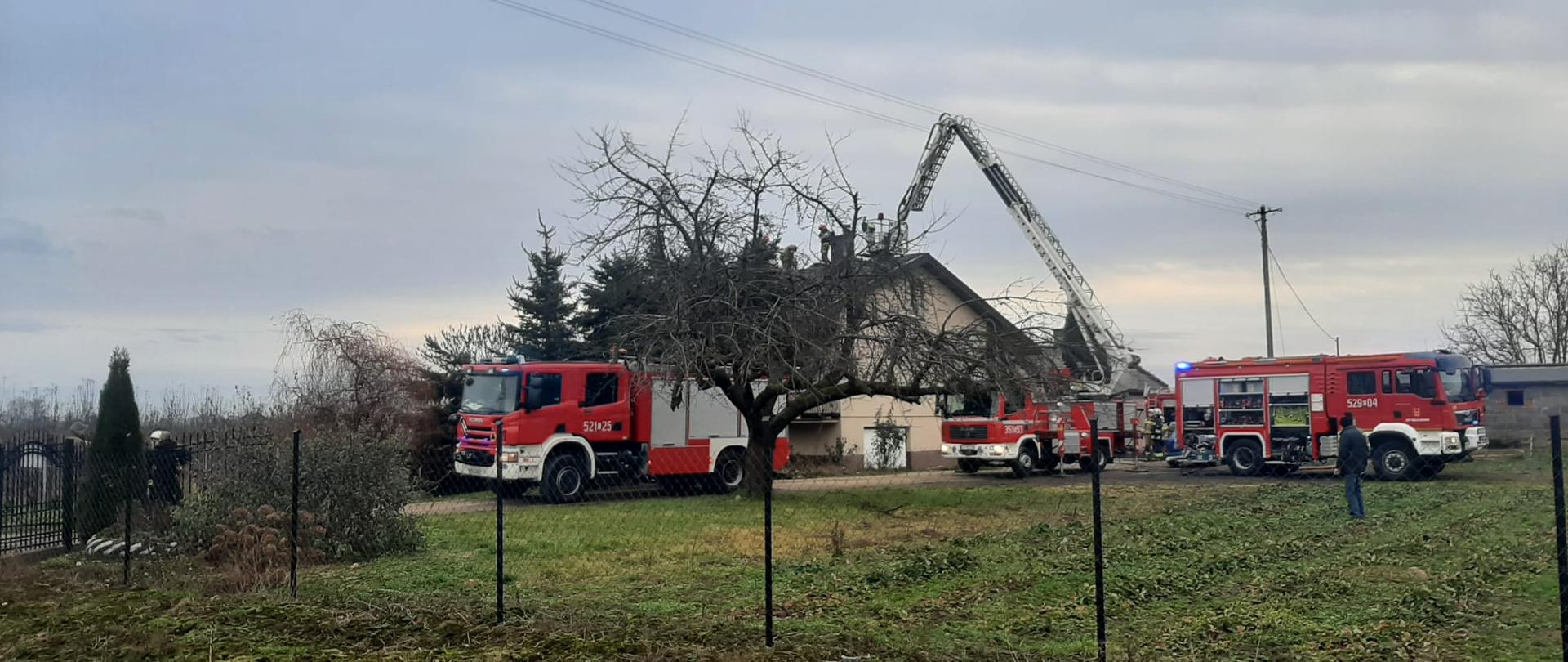Obok domu jednorodzinnego stoją trzy samochody strażackie, z czego jeden to podnośnik strażacki, który ma rozłożoną drabinę w kierunku dachu domu. Na dachu znajdują się strażacy, który dogaszają pożar dachu domu jednorodzinnego.
