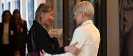 Two women are greeting each other, smiling and touching each other's forearms