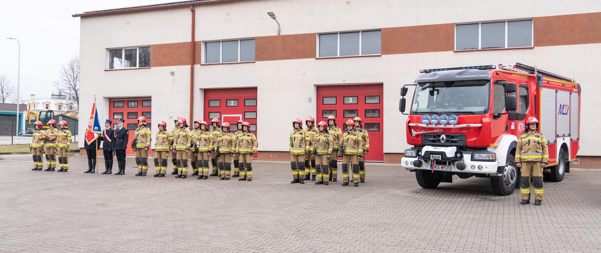 Zdjęcie zrobione na placu wewnętrznym JRG. W tle widać budynek Krajowej Bazy Sił i Środków z czerwonymi bramami. Strażacy ustawieni w szeregu, ubrani w piaskowy strój bojowy oraz czerwony hełm. Z prawej strony stoi nowy czerwony wóz bojowy.